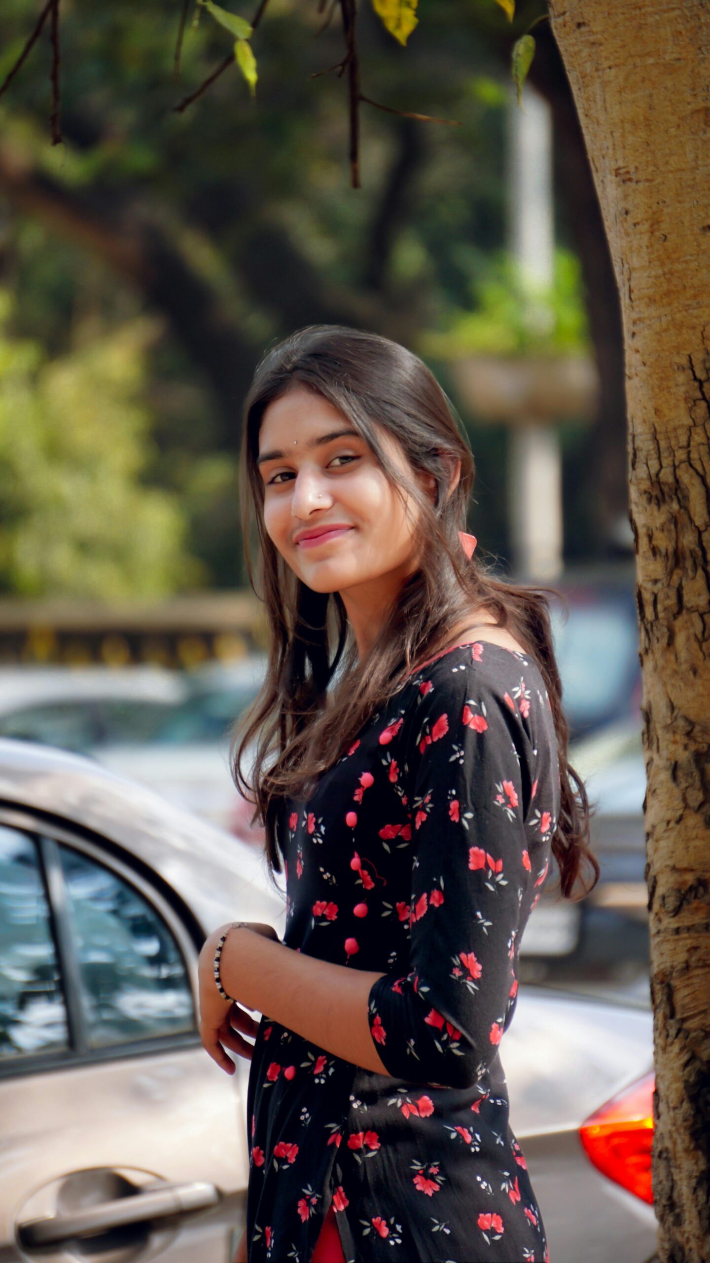 A young woman wearing a floral dress smiles by a tree on a sunny day.
